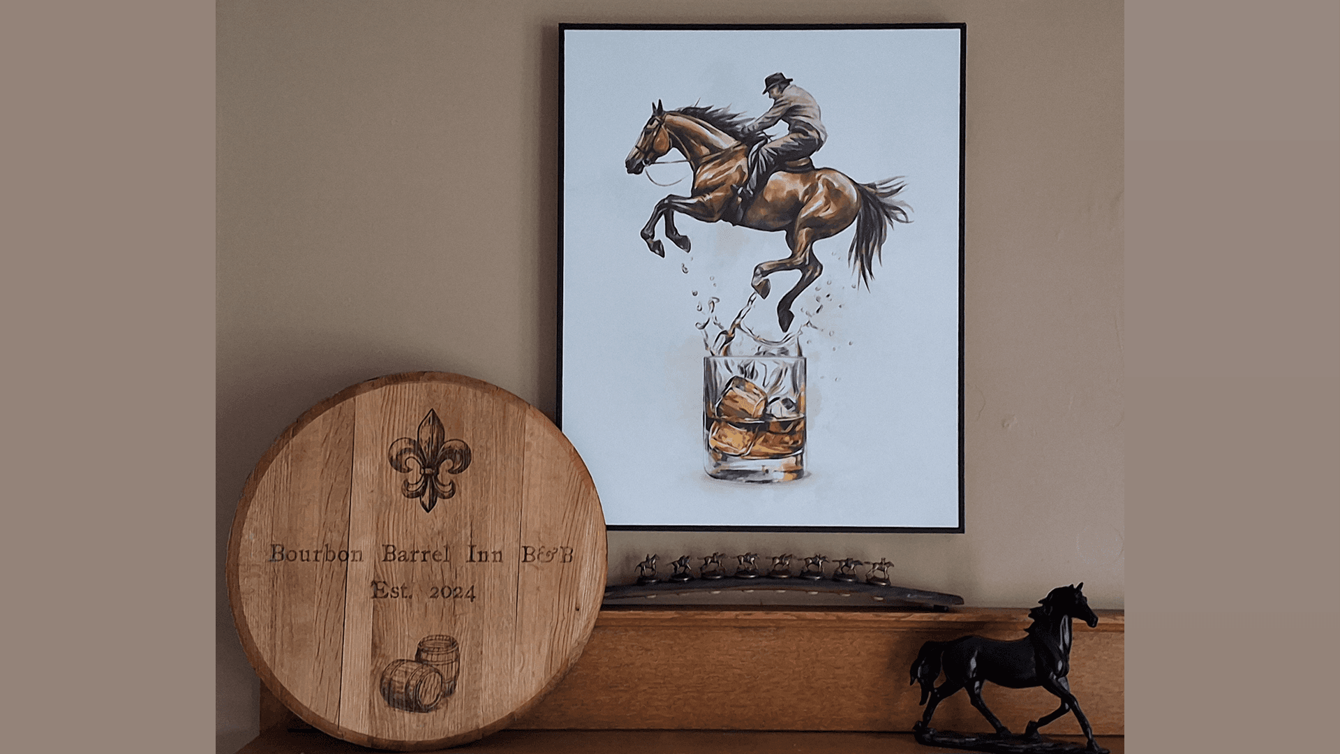 A framed artwork of a horse and rider jumping over a glass of whiskey is displayed on a shelf next to a wooden barrel sign for "Bourbon Barrel Inn."